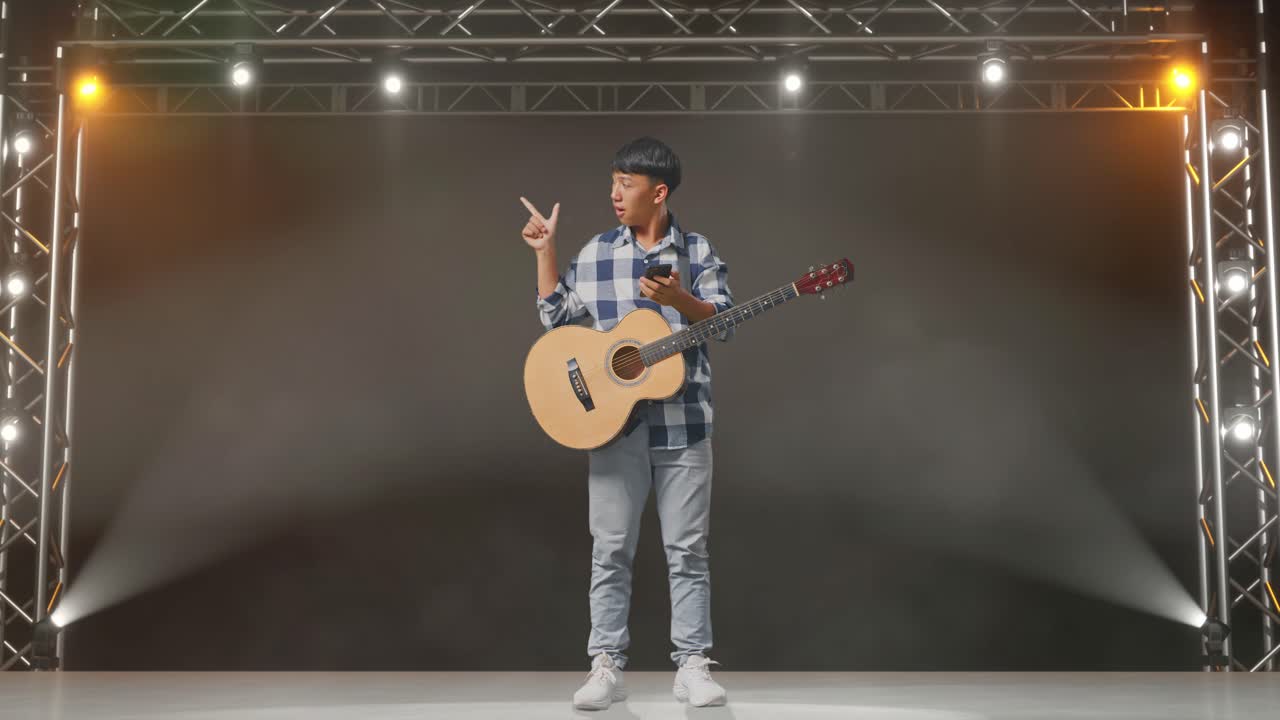 cuerpo lleno de adolescente asiático con guitarra usando teléfono inteligente y señalando al lado mientras está de pie en el escenario