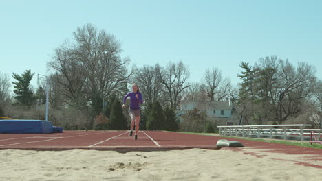 Teen-girl-runs-towards-camera-in-slow-motion-and-jumps-into-a-sand-pit-at-a-track