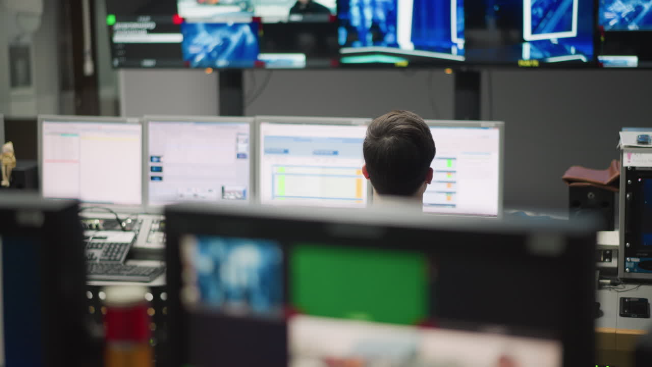 Person monitoring technical production in control room, surrounded by multiple computer screens displaying system data. Operator focused on tasks, managing broadcast systems with multiple controls