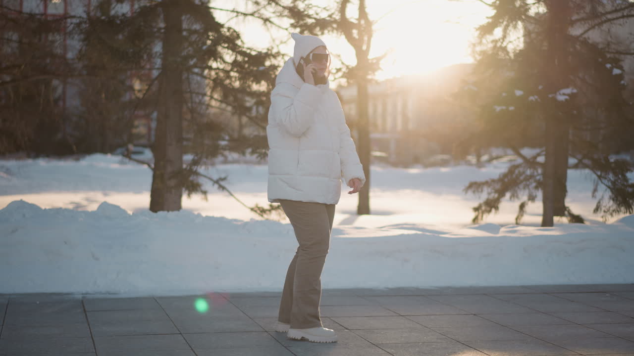 Side view student holds phone to ear singing and dancing in rhythm under warm sunset glow wearing beanie and puffer coat with blurred snowy park and trees conveying joyful winter vibe