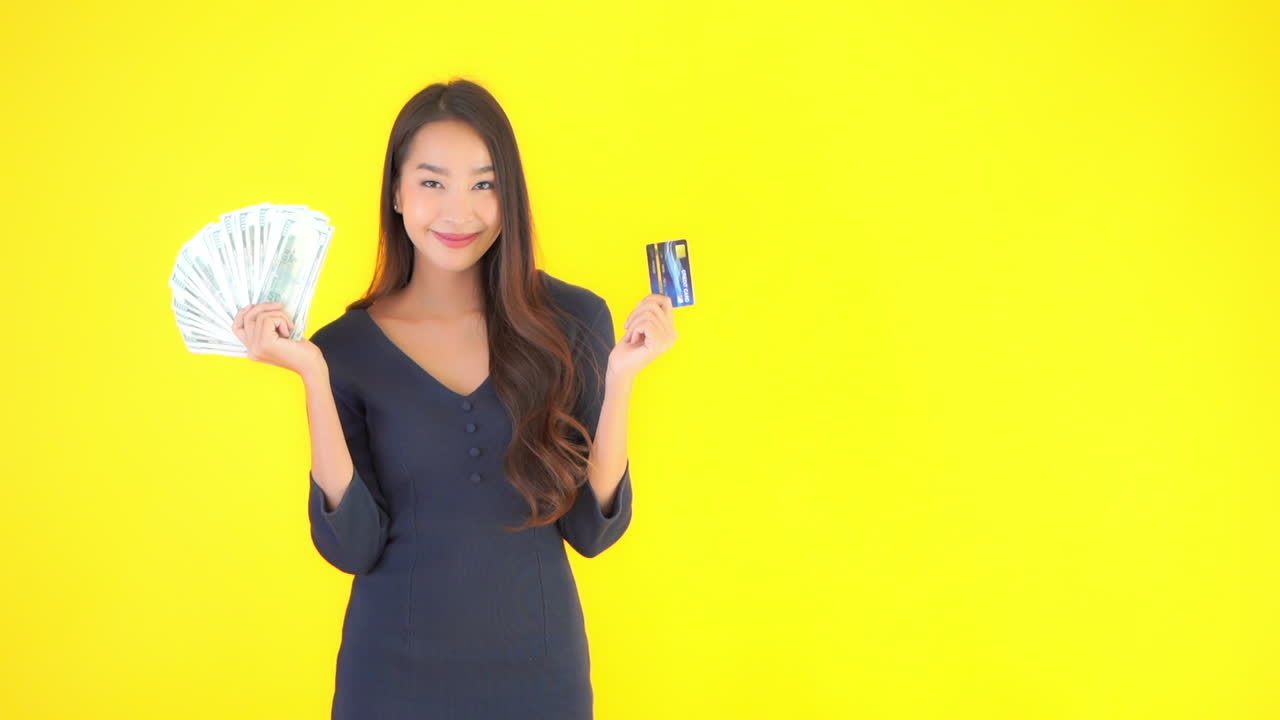 Beautiful Happy Asian Female Holding Cash Dollar Money Bills and Credit Card. Isolated on Yellow Screen Background
