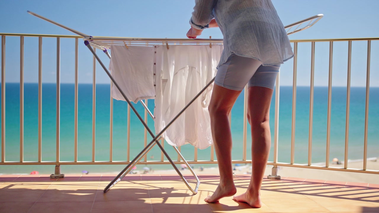 Mulher estendendo roupas brancas em um varal na varanda com vista para o mar durante férias de verão ensolaradas.