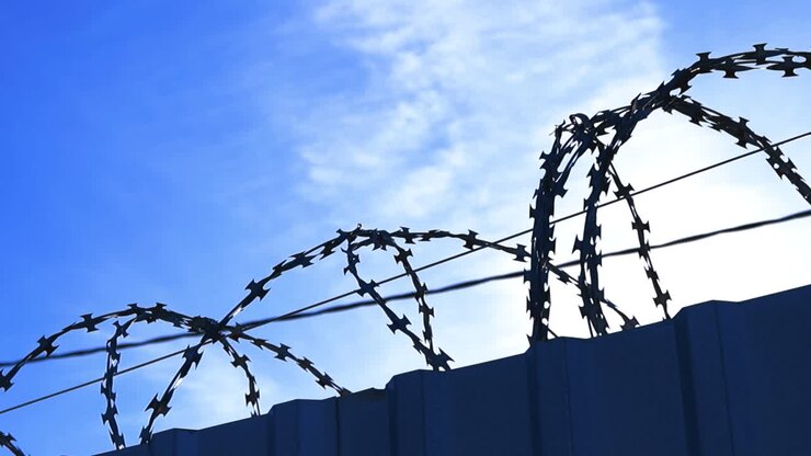 Coils of sharp barbed razor wire sitting atop a high security fence on a sunny day with blue sky and light clouds, symbolizing restriction, confinement, and forbidden access to a private zone