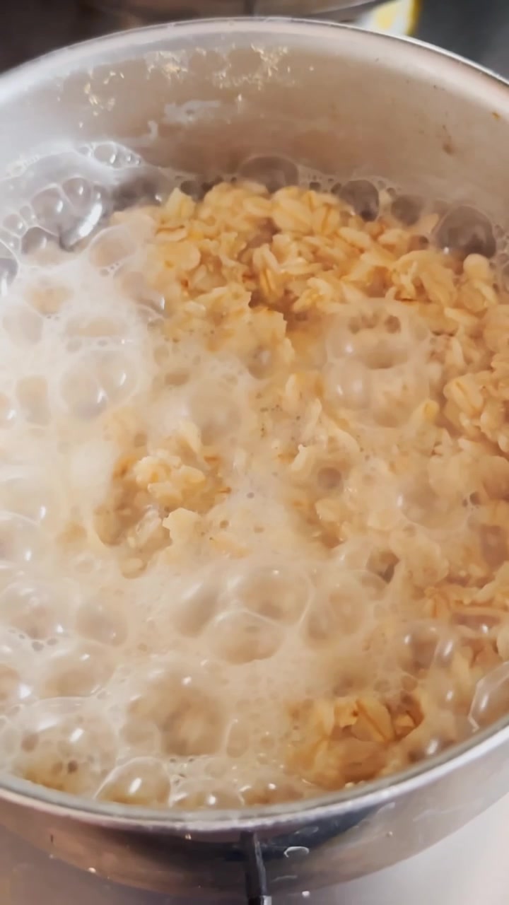Primo piano verticale di porridge in ebollizione in una pentola di metallo, con fiocchi d'avena e bolle cremose che salgono durante la preparazione di una semplice colazione fatta in casa.