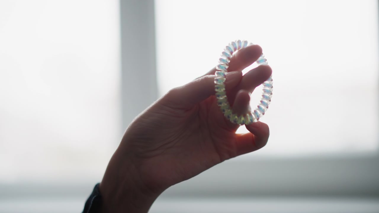 Close up of hand holding and gently rotating iridescent coiled hair tie against soft natural light. translucent plastic object reflects colors as it moves with background blurred