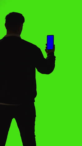 Vertikale-Videoaufnahme-Eines-Mannes,-Der-Ein-Mobiltelefon-Mit-Blauem-Bildschirm-Hält-Und-Von-Hinten-Als-Silhouette-Vor-Einem-Grünen-Bildschirm-Steht