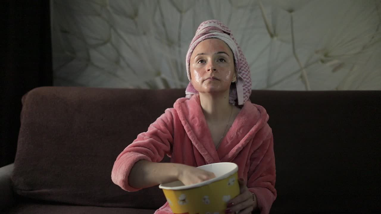 Televizyonda geç saatte film izleyen, patlamış mısır yiyen kadın. Bornoz, yüz maskesi