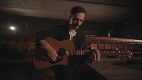 a handsome guy plays an acoustic guitar in an abandoned cinema. the musician sings a song and accompanies on the guitar