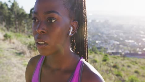 African-american-woman-exercising-outdoors-putting-on-wireless-earphone-in-countryside-at-sunset