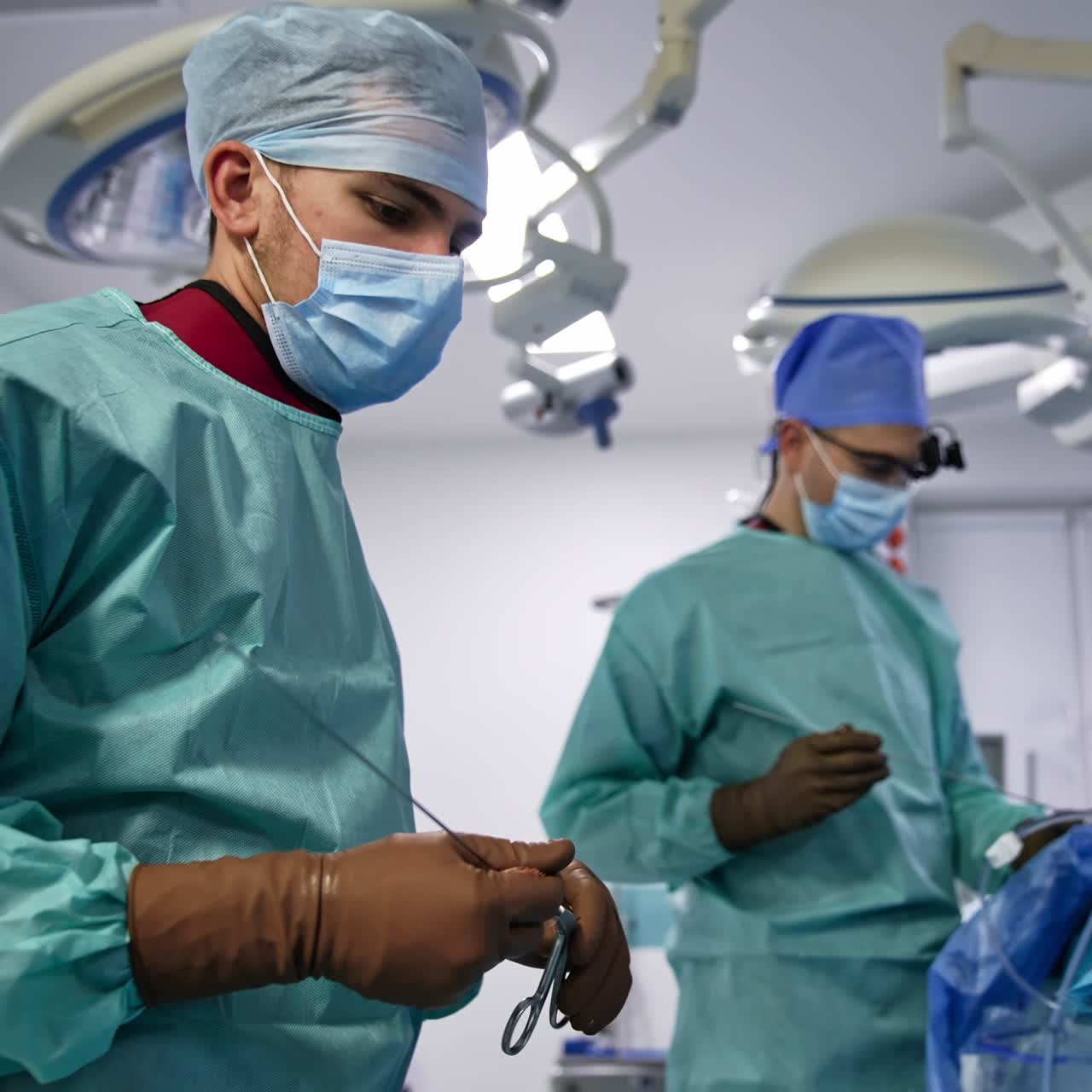 Male assistant passes the long metal needle to the chief surgeon. Doctor at backdrop performs the operation in blur