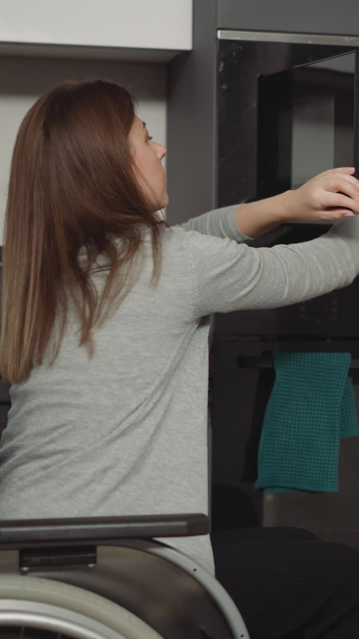 mulher em cadeira de rodas tira um prato com comida aquecida do micro-ondas em casa. jovem mãe sorri carinhosamente olhando para entrar na cozinha filho pequeno