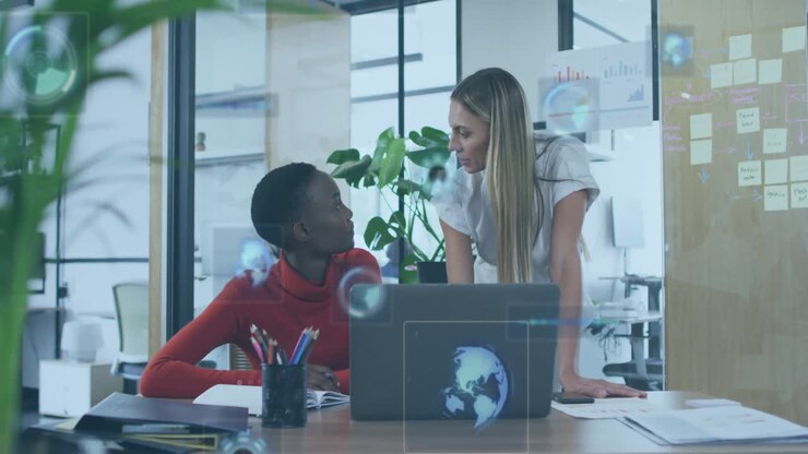 To kvinder diskuterer arbejde ved et kontorbord med animation af en digital globus overlejret.
