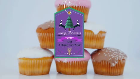 Animation-of-christmas-greetings-on-tag-over-cupcakes-on-white-background