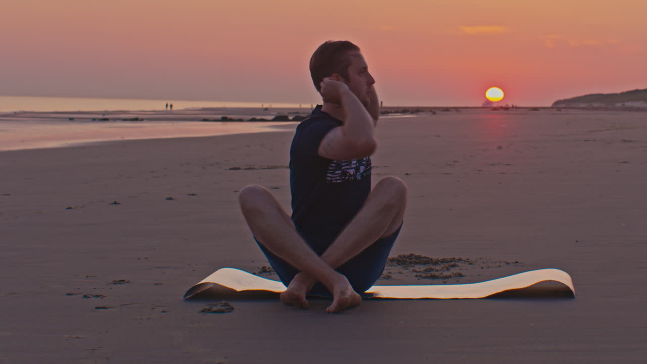 white slim caucasian european Dutch man male model sits on the beach with sunset next to the seashore in Netherlands North sea with setting sun on the background