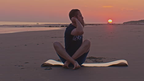 white slim caucasian european dutch man male model sits on the beach with sunset next to the seashore in netherlands north sea with setting sun on the background