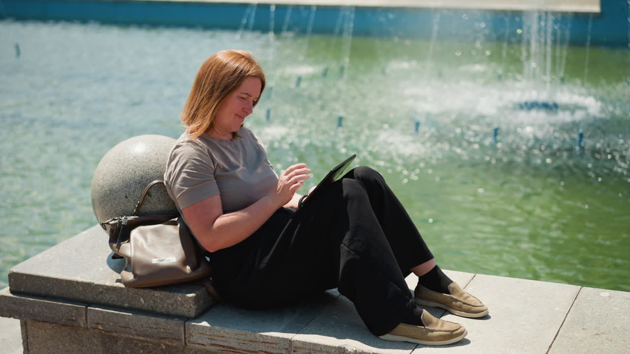 Emotional lady sitting beside fountain operating phone under warm sunlight, relaxed posture showing tenderness and reflection, soft breeze moving hair, tranquil outdoor mood filled with warmth