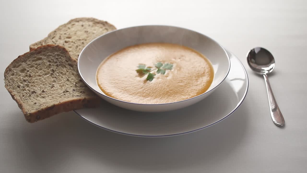 Steaming hot butternut squash soup with a sprig of parsley floating on the surface, in a white bowl on a white table with sliced bread and a spoon at the side.