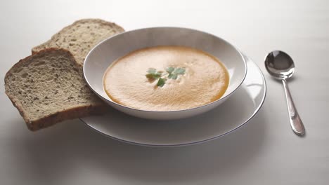 steaming hot butternut squash soup with a sprig of parsley floating on the surface, in a white bowl on a white table with sliced bread and a spoon at the side.
