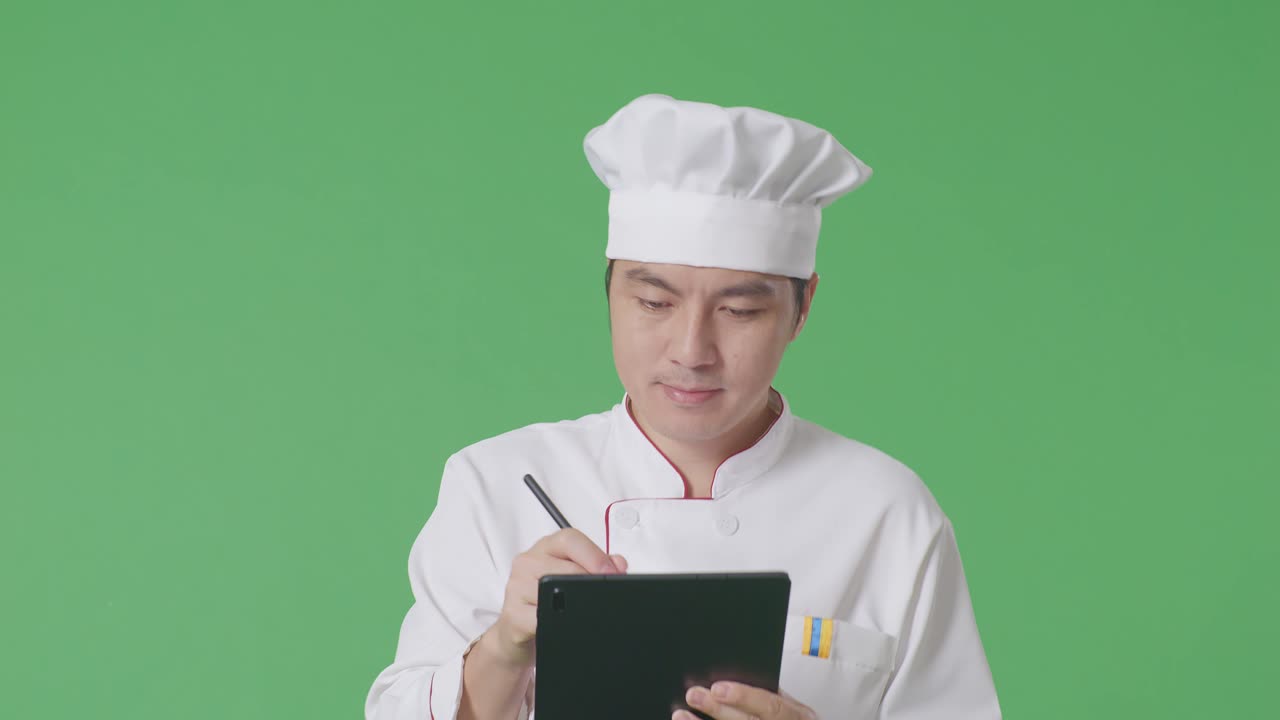 Chef asiatique homme prenant des notes sur une tablette en marchant dans un studio sur fond vert