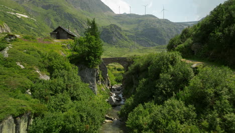 Green-meadow-with-lots-of-vegetation-and-shot-under-an-old-stone-bridge,-wind-power-windmills-on-the-horizon