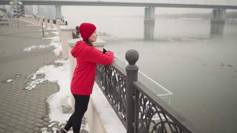 woman in red hoodie and black leggings resting hands on decorative metal railing overlooking calm river during winter, snow-covered ground, distant bridge with moving vehicles