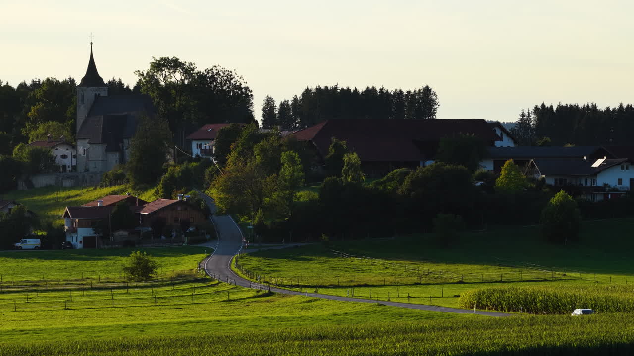 Cinematic aerial drone shot of a small rural village with church and winding road surrounded by green farmland in the European alpine countryside during golden hour