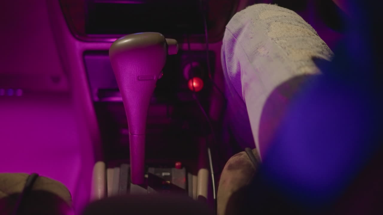 Interni auto di notte, mano sulla leva del cambio, luce viola neon sul ginocchio in denim, luci del cruscotto e console centrale illuminate, primo piano del conducente che raggiunge la console, atmosfera cinematografica al rallentatore, tranquilla area urbana