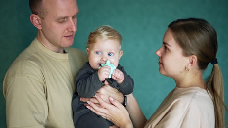 caucasian parents with a little chubby baby boy. cute infant child chewing a toy. blue backdrop.