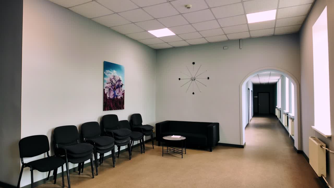 Minimalist medical waiting room: stacked black chairs, abstract flower artwork, radial wall clock and bright archway leading to exam offices. Daugavpils, Latvia (Daugavpils, Latvija)