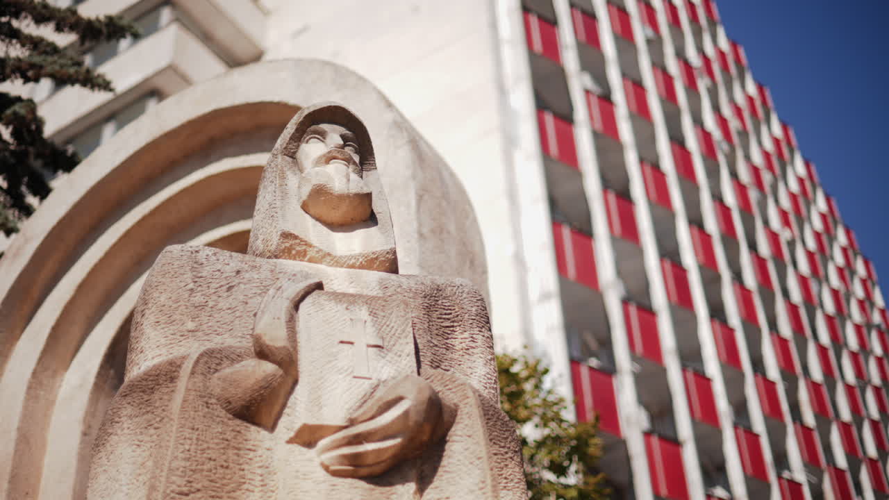 Low angle view of a carved stone statue Petru Movila holding a book with a cross, framed by trees and modern architecture, Chisinau, Moldova