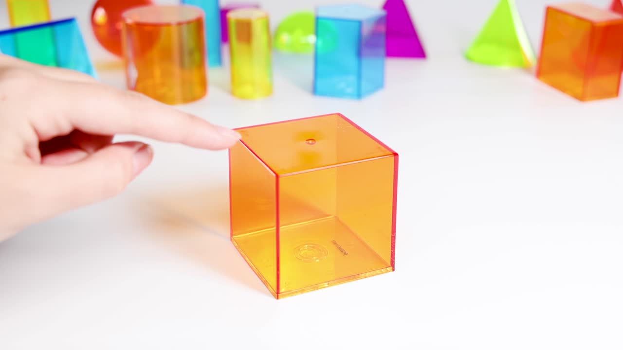 Hands manipulate vibrant 3D shapes on a white surface, exploring geometry with colorful prisms and cubes
