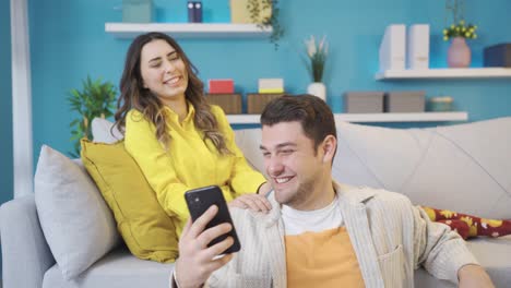 Happy-couple-sitting-on-sofa-at-home-chatting,-looking-at-phone-and-laughing.