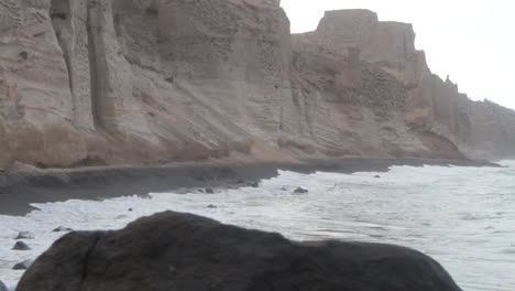 Kamerafahrt-Von-Wellen,-Die-Auf-Einen-Schwarzen-Strand-Treffen,-Der-Von-Hohen-Weißen-Vulkanischen-Klippen-Umgeben-Ist