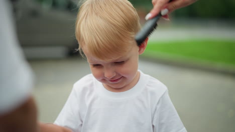 a happy little boy smiles brightly as a woman combs his hair, dressed in a white top, he stands outdoors on a paved path with a back view of another boy in white