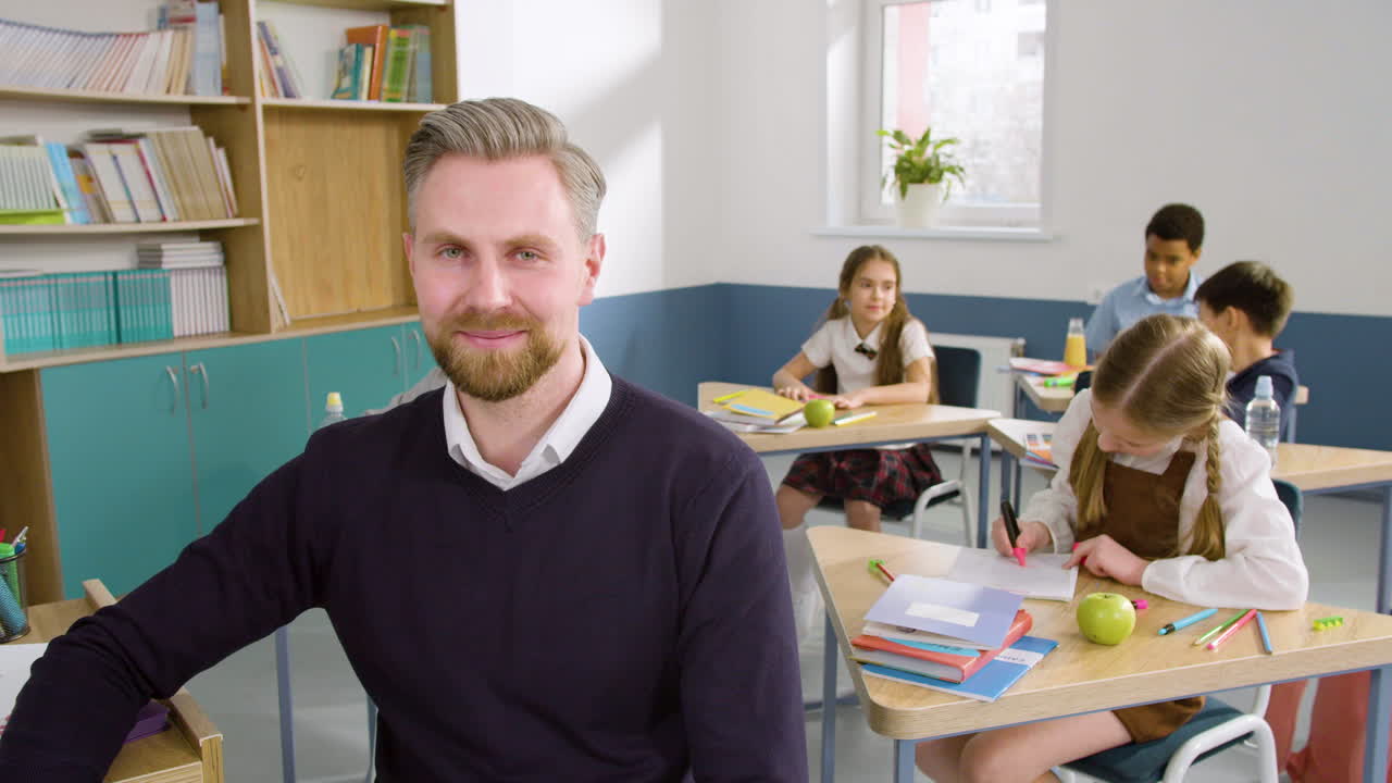 profesor mirando la cámara en el aula de inglés, en el fondo sus estudiantes están sentados en los escritorios