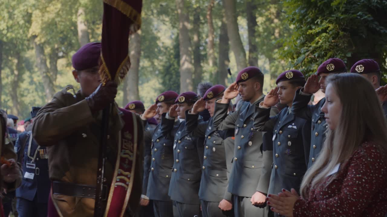 Soldiers from a paratrooper unit salute and hold ceremonial flags during a solemn military commemoration event. The scene captures respect and honor in a formal setting. Airborne, Oosterbeek, Holland