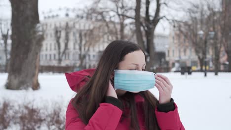 caucasian woman in red hooded coat puts on medical mask in winter park