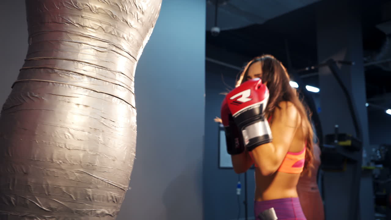 Energetic woman wearing gloves in the gym boxing big pear and articulates something with her mouth. Concentrated fighter girl confidently punches a pear