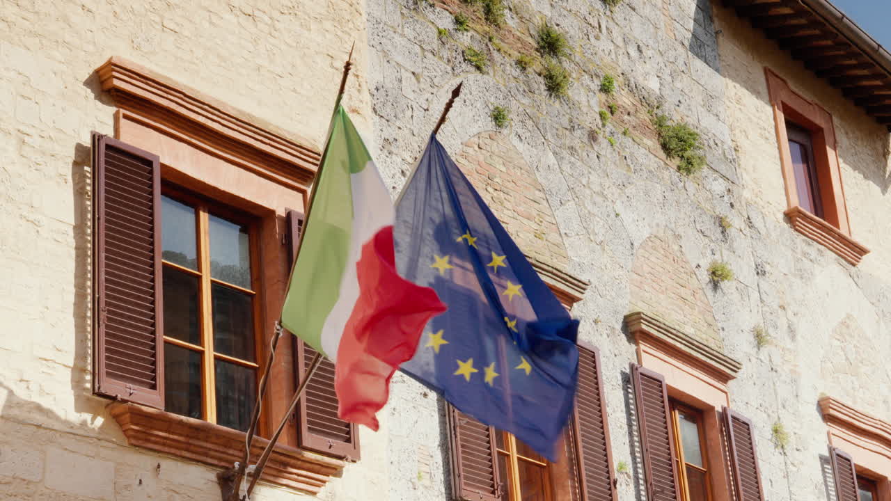 Bandiere italiana e dell'UE che sventolano su un edificio rustico toscano.