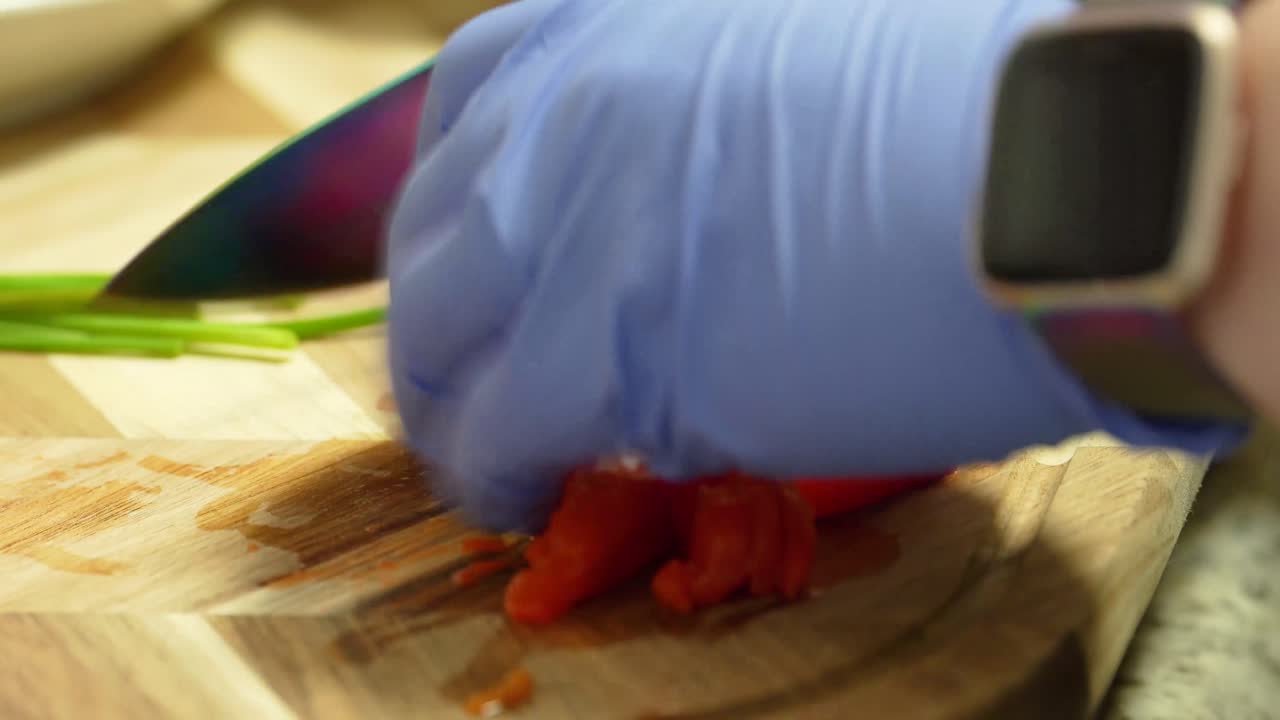 Chef Wearing Blue Rubber Gloves Chops and Dice Red Peppers on a Cutting Board using a Large Sharp Knife in a Kitchen surrounded by Food in the Background, Closeup no face