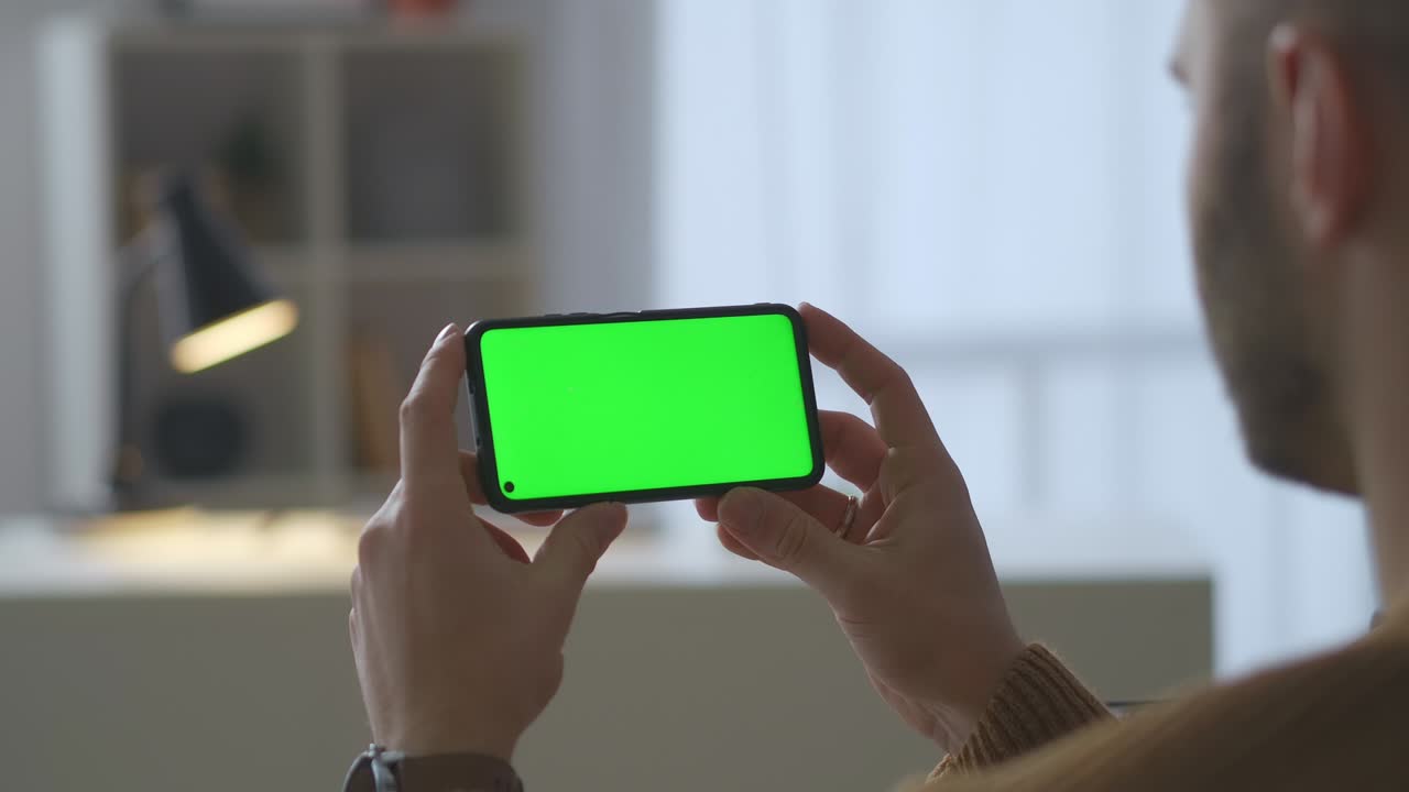 집에 앉아서 비디오를 보는 사람의 손에 있는 현대 스마트폰의 녹색 화면, 어깨를 통해 근접한 시각