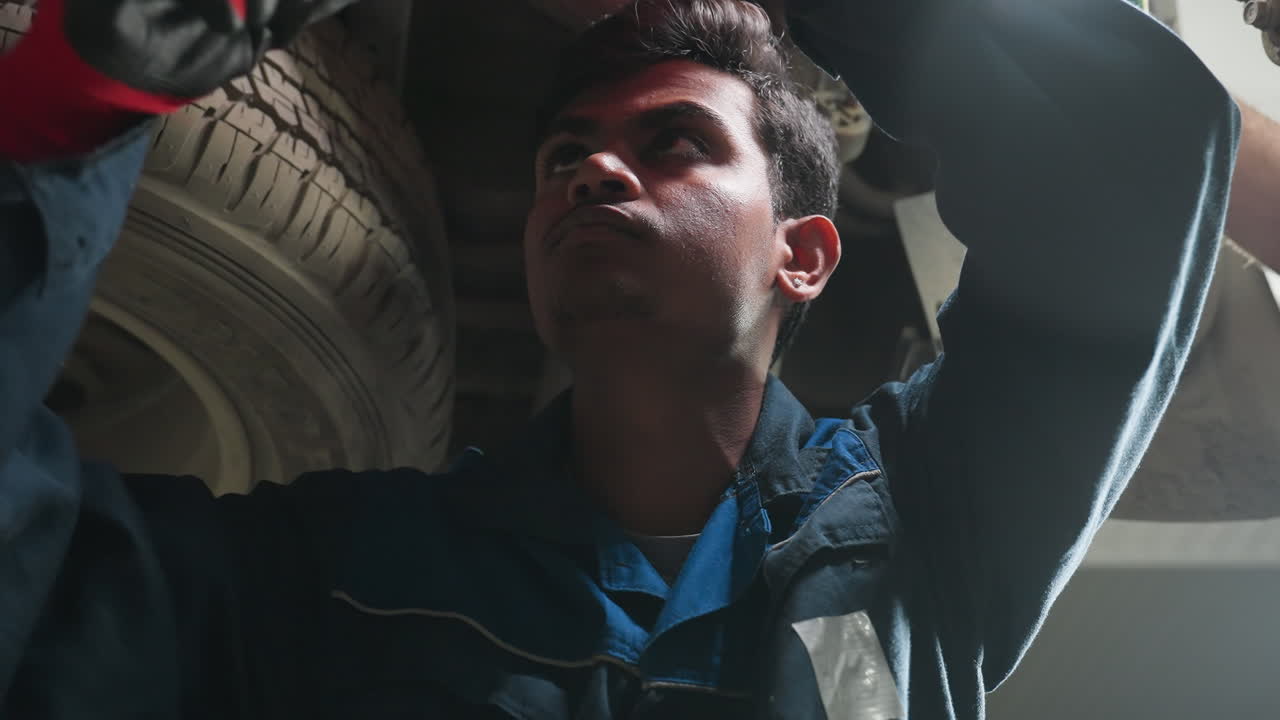 Engineer working under car, tightening bolt with focused expression, light reflecting on face, wearing gloves and blue uniform, concentrated automotive repair in dimly lit workshop