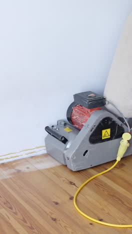 an industrial floor sander is used on pine wood planks near the wall, guided by unrecognizable worker in sneakers during a renovation process, vertical framing, camera static, real time