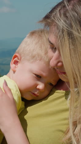 careful smiling mother with long hair calms down sleepy little son stroking back standing against old distant mountains at resort closeup slow motion