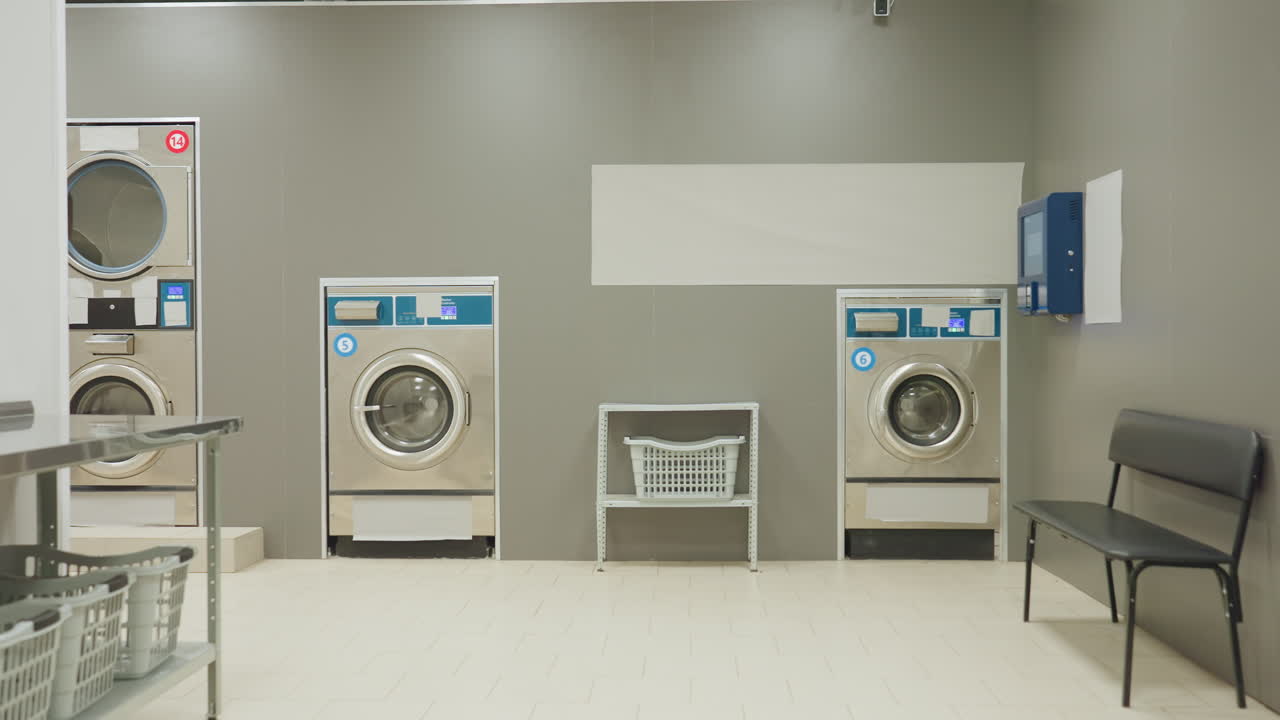Laundry worker inside laundry room turning off washing machine before leaving, walking towards entrance to switch off light, machines, bench, laundry basket visible in service facility environment