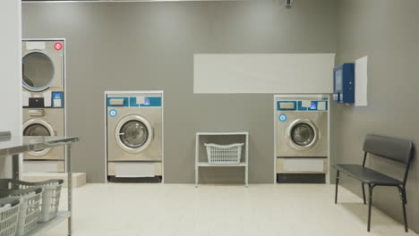 laundry worker inside laundry room turning off washing machine before leaving, walking towards entrance to switch off light, machines, bench, laundry basket visible in service facility environment
