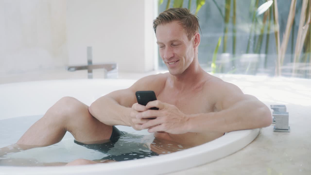 젊은 남자가 현대적인 욕조에서 스마트폰에서 메시지를 읽습니다.