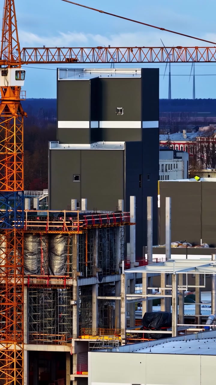 Close-up drone flight over a large industrial construction site in a snow-covered rural town in Northern Europe, with wind turbines in the background, showcasing cold and modern energy development.