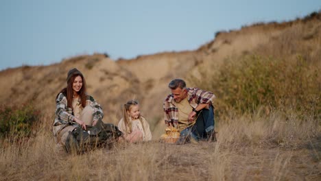 a middle-aged man in a plaid shirt together with his wife, a brunette girl in a green plaid shirt, are relaxing during their picnic outside the city and taking out a thermos to drink tea on a summer evening