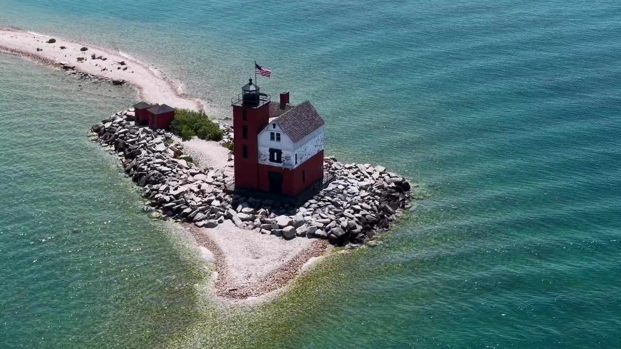 Vista aerea ravvicinata sopra il faro di Round Island e la sua diga frangiflutti in pietra, circondati da acqua verde limpida vicino a Hessel, Michigan.
